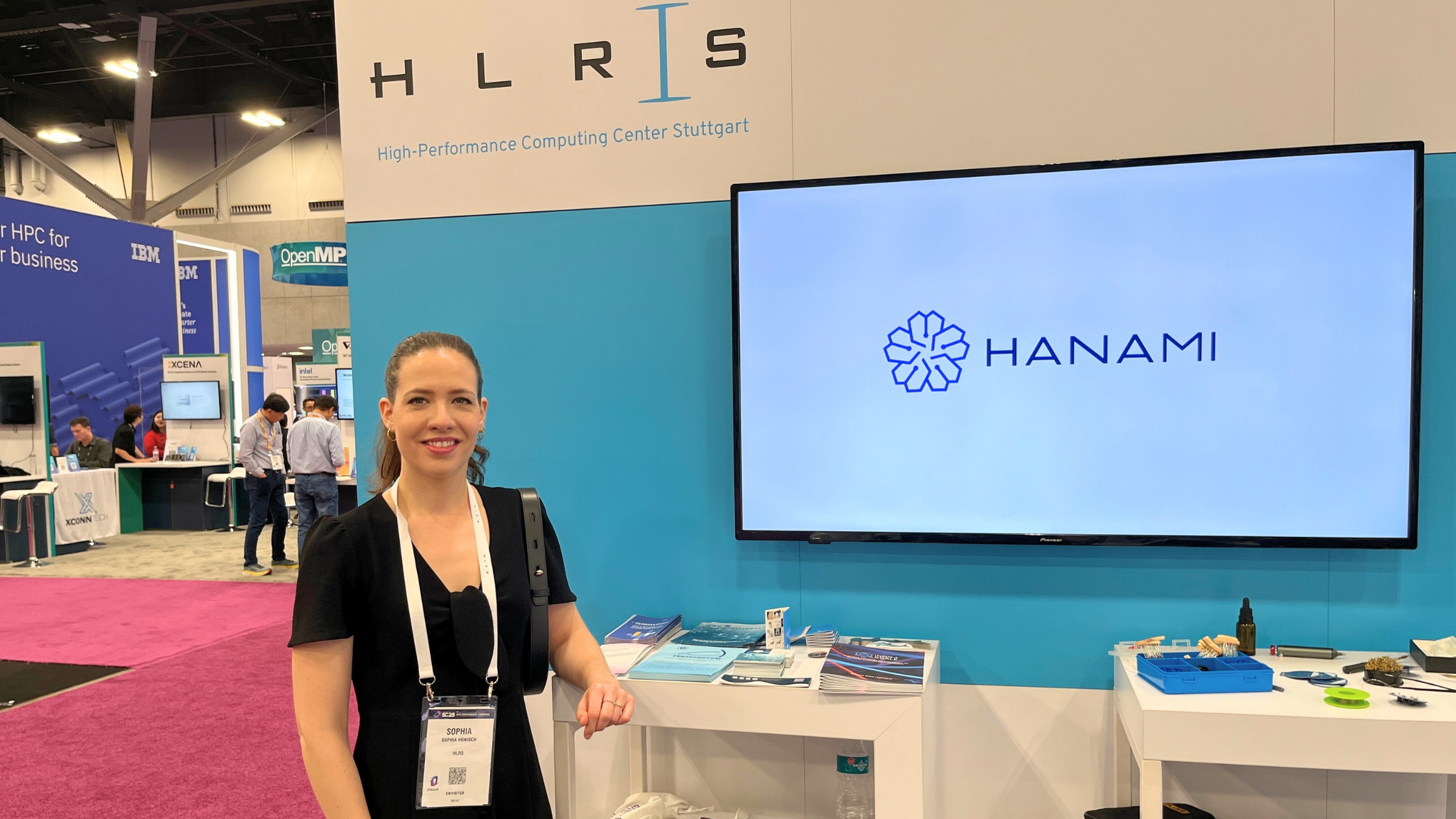 A woman stands at the HANAMI booth during an HPC event, next to a display screen showing the HANAMI logo.