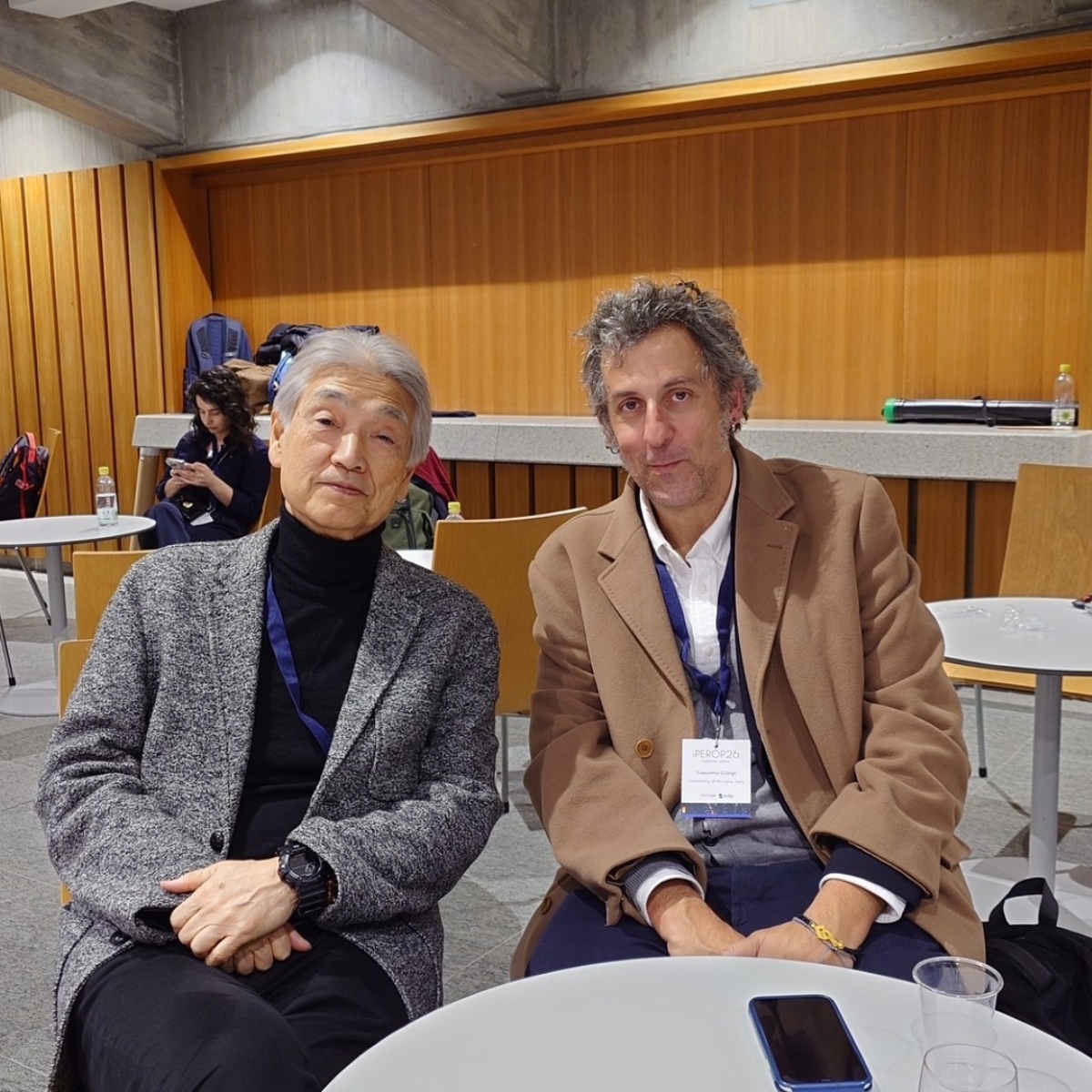 Two man sitting on wooden chairs and posing for the photo.