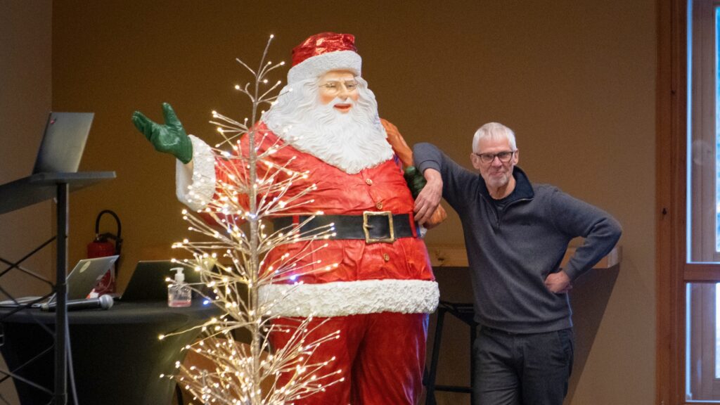 Man posing with a life-sized Santa Claus decoration indoors, standing beside a small illuminated decorative tree, during the HANAMI high-level symposium, in Chamonix.