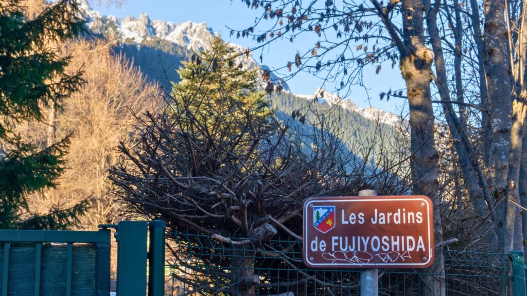 Sign reading “Les Jardins de Fujiyoshida” mounted on a fence, with alpine mountains and trees in the background.