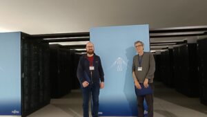 Two researchers standing between rows of supercomputer racks, in front of a blue panel featuring Mount Fuji and Fujitsu branding, inside a high-performance computing facility.