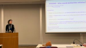 Speaker presenting the HANAMI project during a conference session, standing at a lectern beside a slide titled “HANAMI – Who stands behind this initiative?”, outlining Horizon Europe funding, project timeline, and EU–Japan digital cooperation objectives.