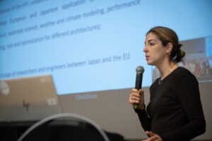 Woman, with a black sweater, talking to a microphone at a conference podium, with slides in the background.