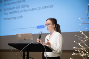 Woman, with a white blouse, talking to a microphone at a conference podium, with slides in the background.