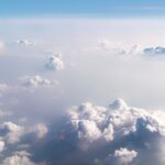 Aerial view of scattered white clouds stretching across the sky, with soft sunlight illuminating the cloud tops above a hazy horizon.
