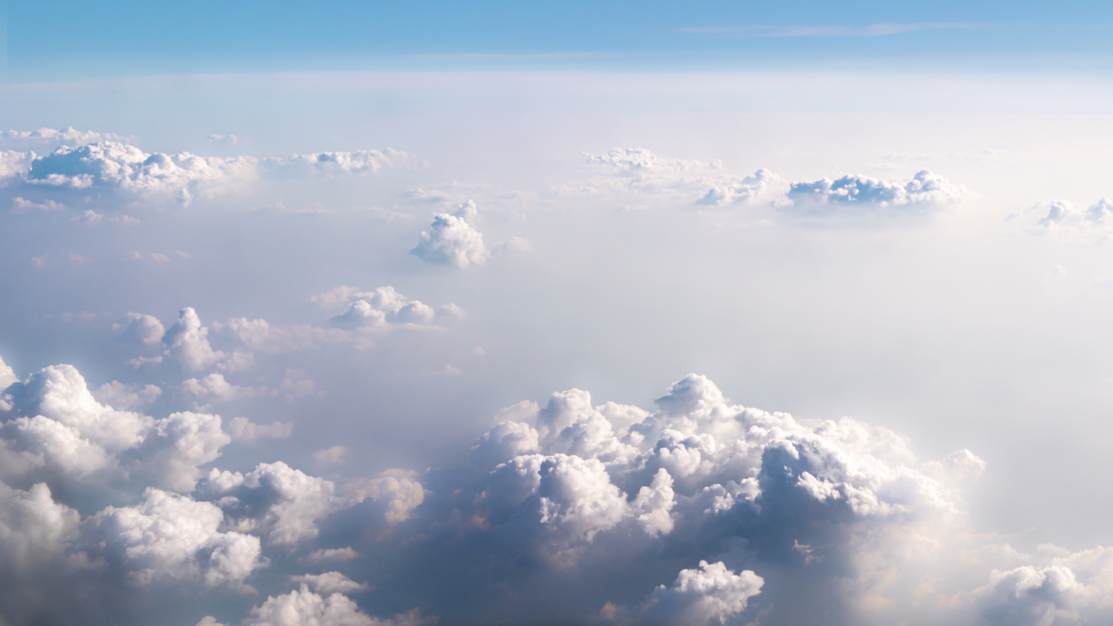 Aerial view of scattered white clouds stretching across the sky, with soft sunlight illuminating the cloud tops above a hazy horizon.